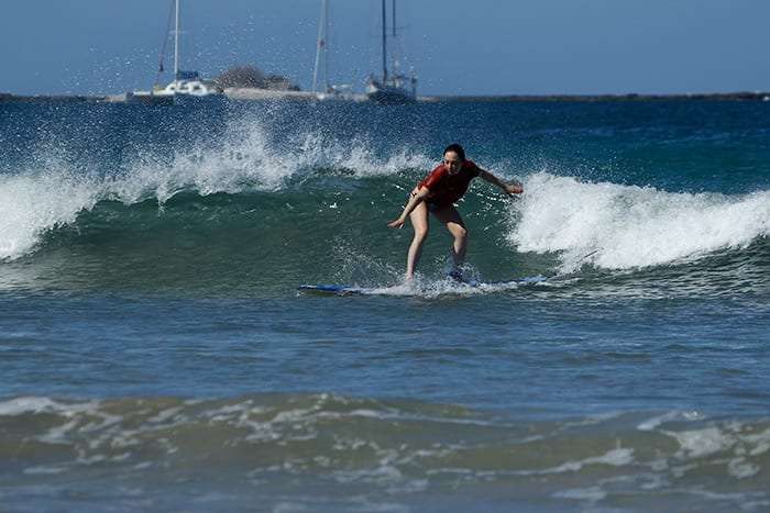 Surfer in a red top riding a small wave with sailboats in the distance