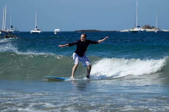Surfer in a black rash guard and plaid shorts riding a wave with boats behind