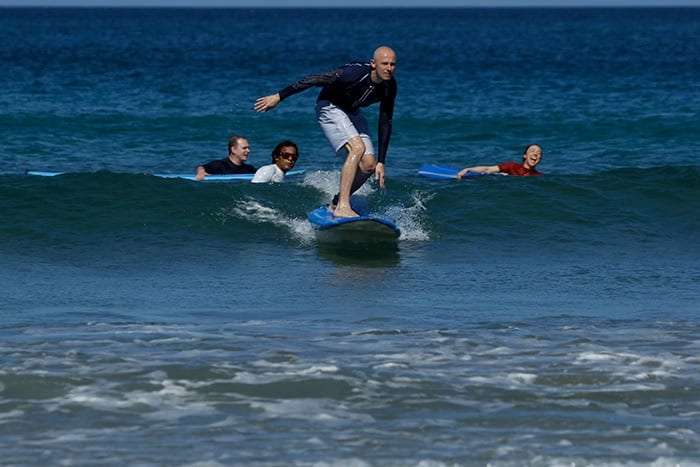 Bald surfer standing on a blue board on a wave while two students paddle behind