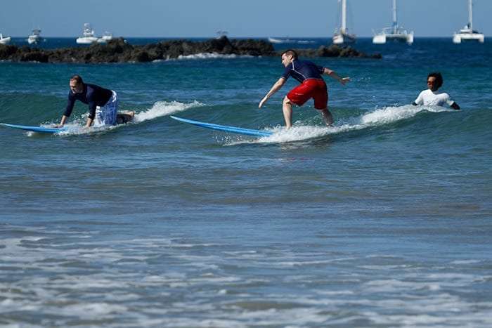 Surf students riding and paddling small waves with anchored sailboats behind