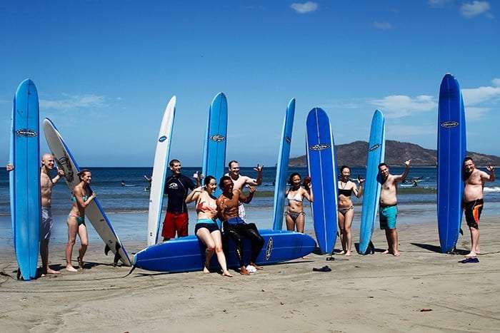 Group of smiling people on a beach with upright blue surfboards