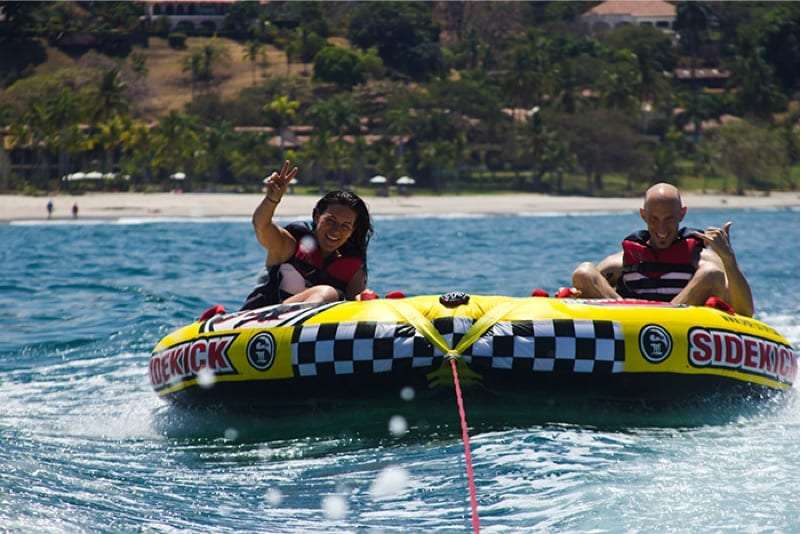 Two people on a yellow inflatable tube pulled behind a boat, one waving