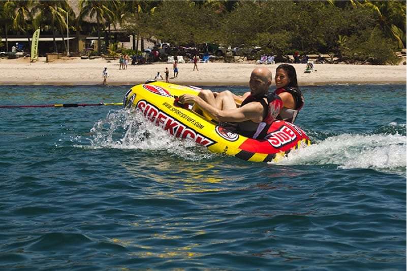 Bald man and a woman riding a yellow and red Sidekick tube towed across the sea off a beach
