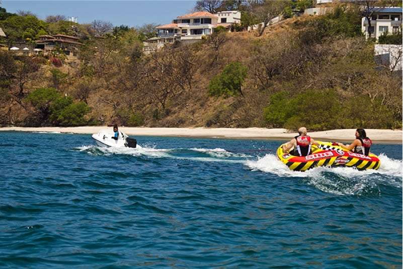 Two people on a yellow and red tube towed behind a small boat along a hilly coast
