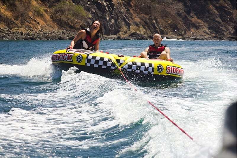 Two riders laughing on a yellow checkered tube bouncing over waves along a rocky shore