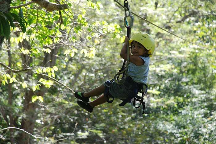 Child in a yellow helmet and harness riding a zip line through the trees