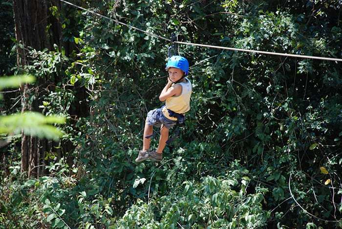 Child in a blue helmet riding a zip line through green forest
