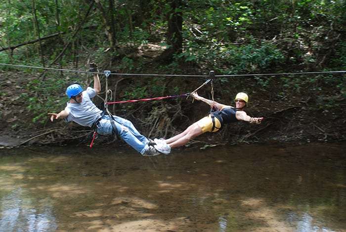 Man and woman in helmets zip lining side by side over a shallow creek