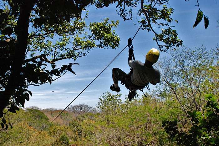 Zip line rider in a yellow helmet seen from below against blue sky and treetops