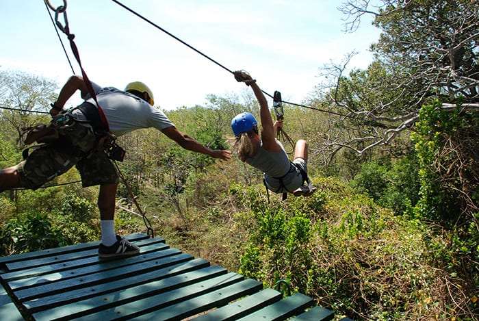 Woman in a blue helmet leaving a wooden platform on a zip line as a guide reaches out