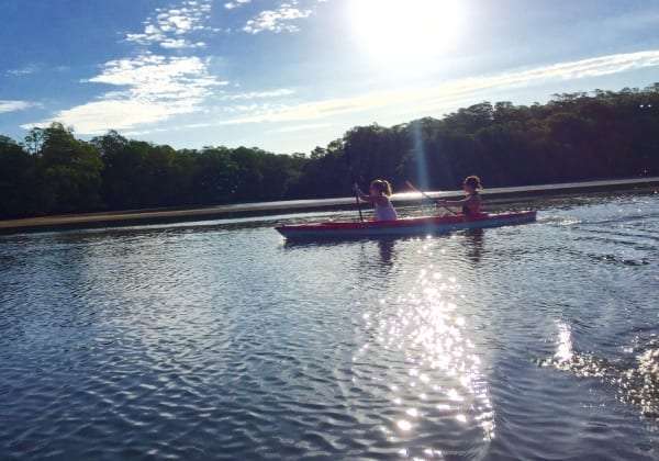Two kayakers paddling a red kayak across calm water at sunset beside a tree-lined shore