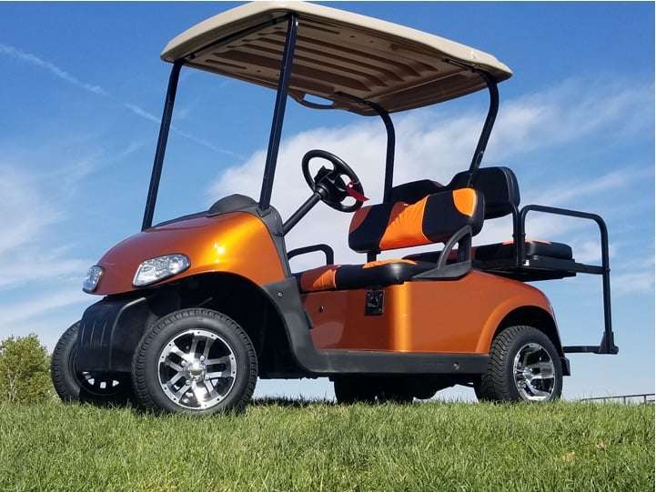 Orange golf cart with a beige roof and a rear flip seat parked on a grass lawn under blue sky