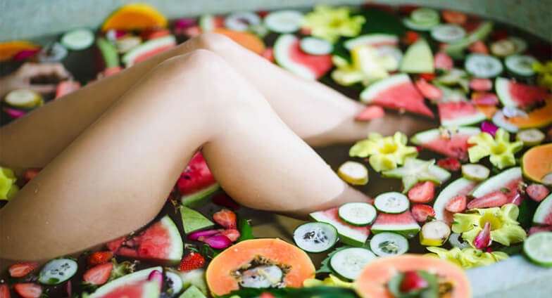 Bath filled with fruit slices and tropical flowers around a woman's bent legs
