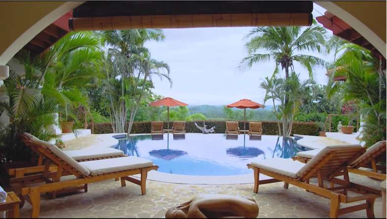Infinity pool seen between wooden sun loungers with palm trees and forested hills beyond