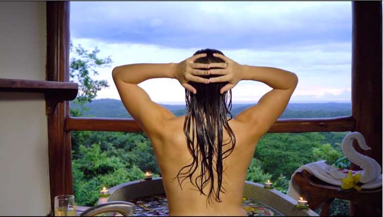 Woman from behind washing her hair in an outdoor tub with a view over green hills